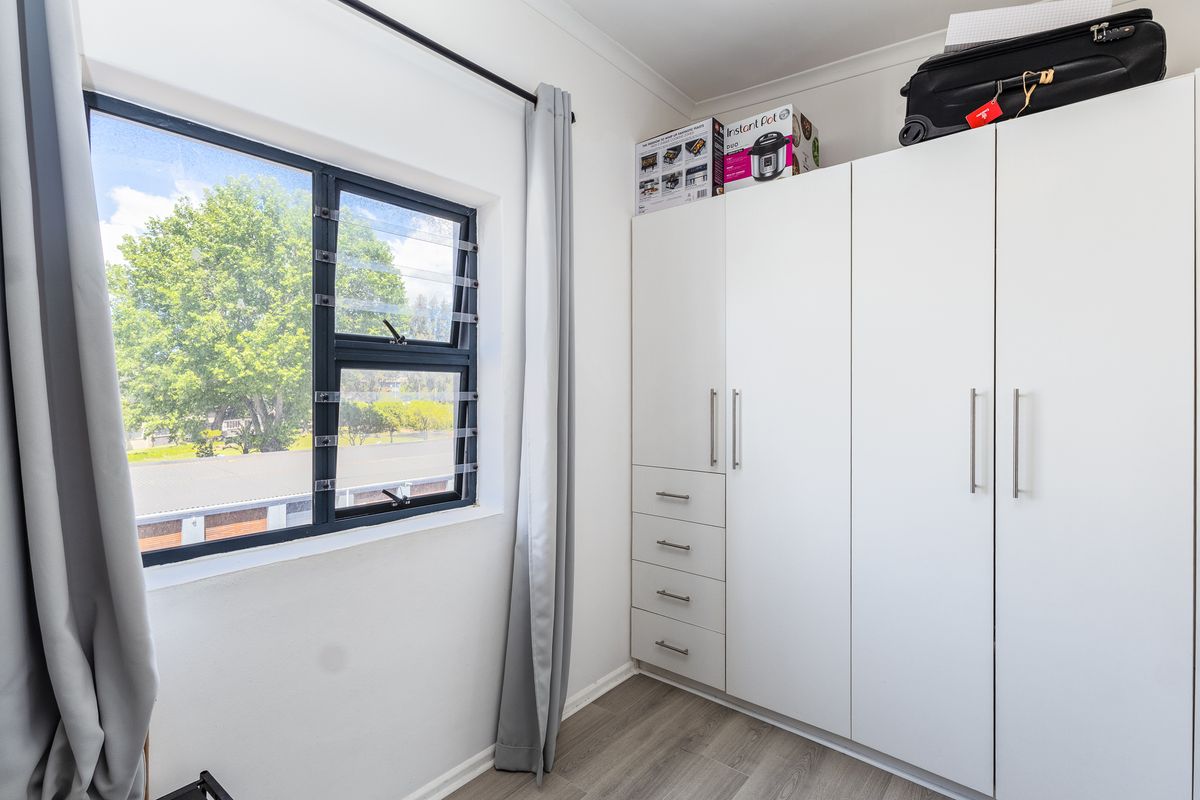 A bright bedroom featuring modern white built-in cupboards, laminate flooring, and a large window providing natural light.