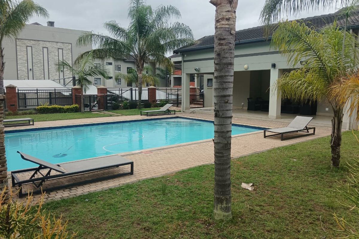 A well-maintained communal swimming pool area featuring a paved deck, surrounding garden, and a covered patio space for outdoor relaxation.