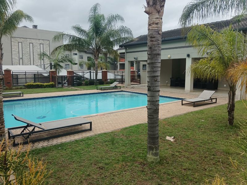 A well-maintained communal swimming pool area featuring a paved deck, surrounding garden, and a covered patio space for outdoor relaxation.