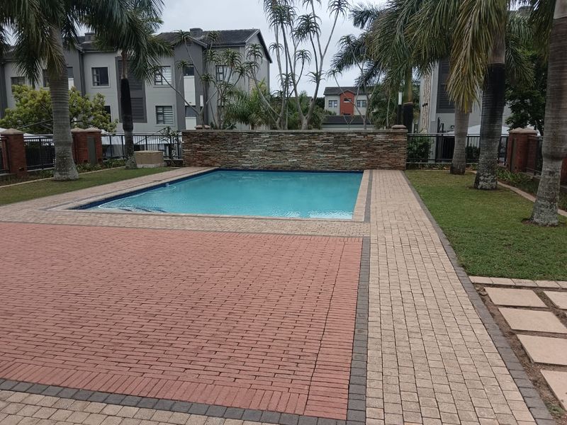 A well-maintained communal swimming pool area featuring a stone-clad feature wall, paved decking, and established palm trees within a residential apartment complex.