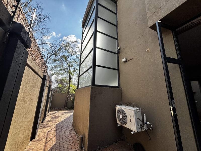 A rear exterior view of the property featuring a paved walkway, boundary wall with electric fencing, and a prominent vertical glass window feature.
