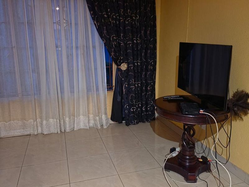 A living room area featuring neutral tiled flooring, recessed ceiling lighting, and large windows dressed with layered curtains.