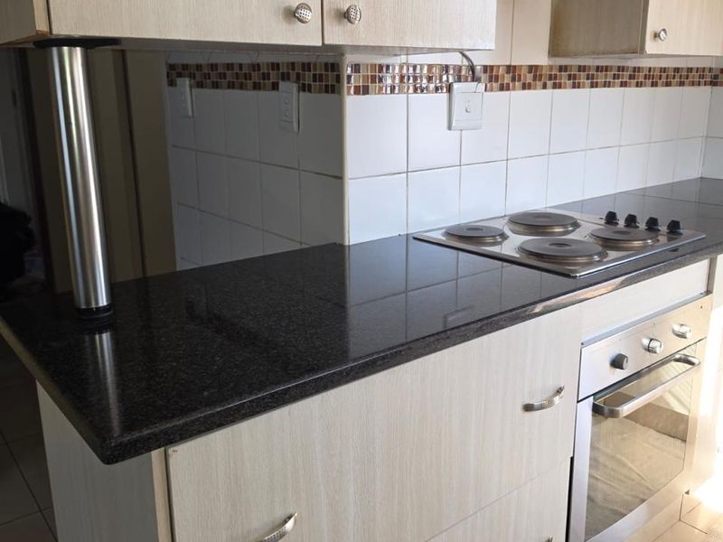 A functional kitchen featuring light wood-finish cabinetry, dark granite countertops, a built-in electric hob, oven, and a tiled backsplash.