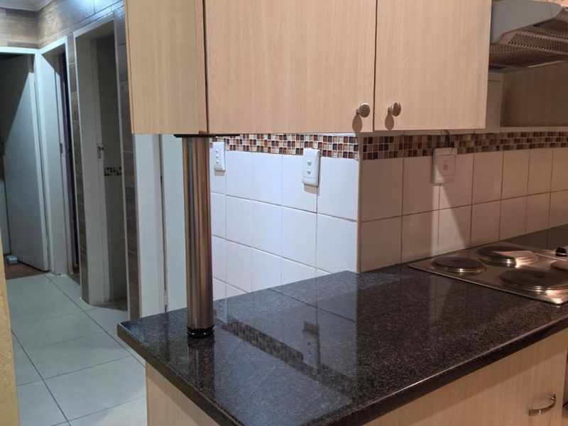 A functional kitchen area featuring light wood-look cabinetry, dark granite countertops, a built-in electric hob with extractor fan, and neutral tiled flooring.