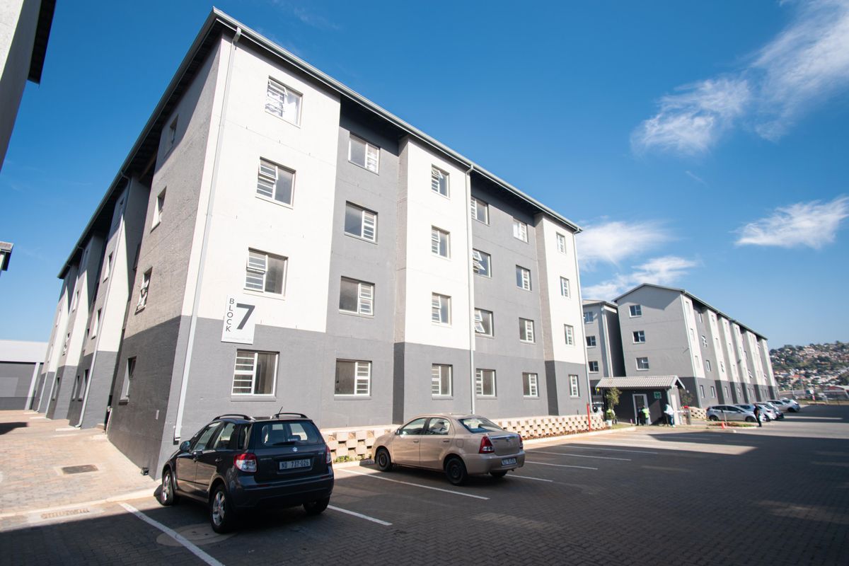 A view of the exterior facade of a multi-story apartment block with designated paved parking spaces in front.