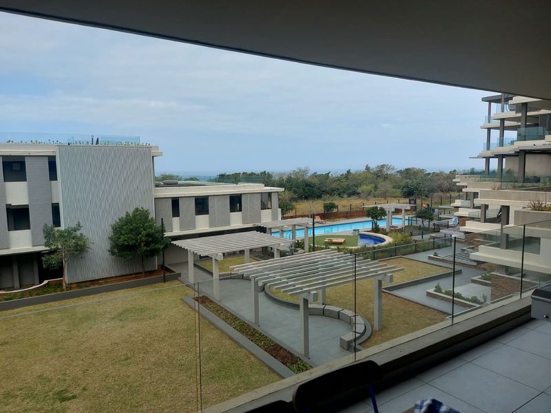 Elevated view from a balcony overlooking a modern apartment complex courtyard, featuring a swimming pool, landscaped gardens, and pergola structures.