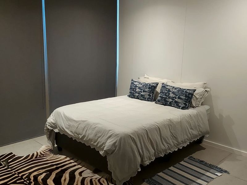 A modern bedroom featuring neutral tiled flooring, recessed downlighting, and dark roller blinds for privacy.
