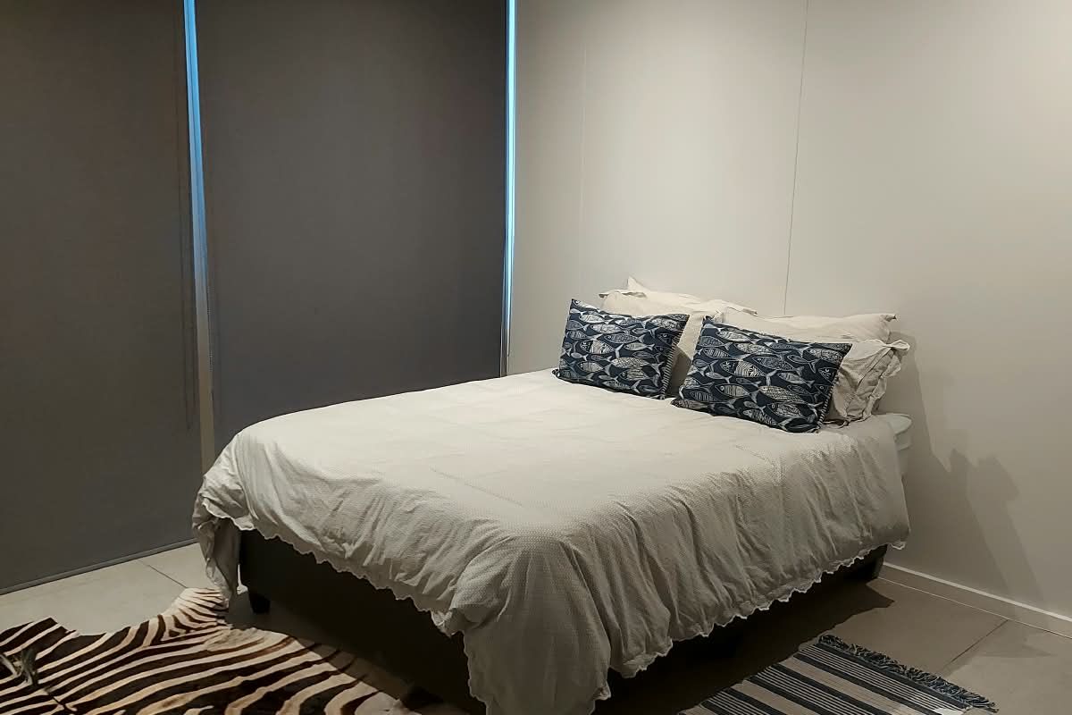 A modern bedroom featuring neutral tiled flooring, recessed downlighting, and dark roller blinds for privacy.