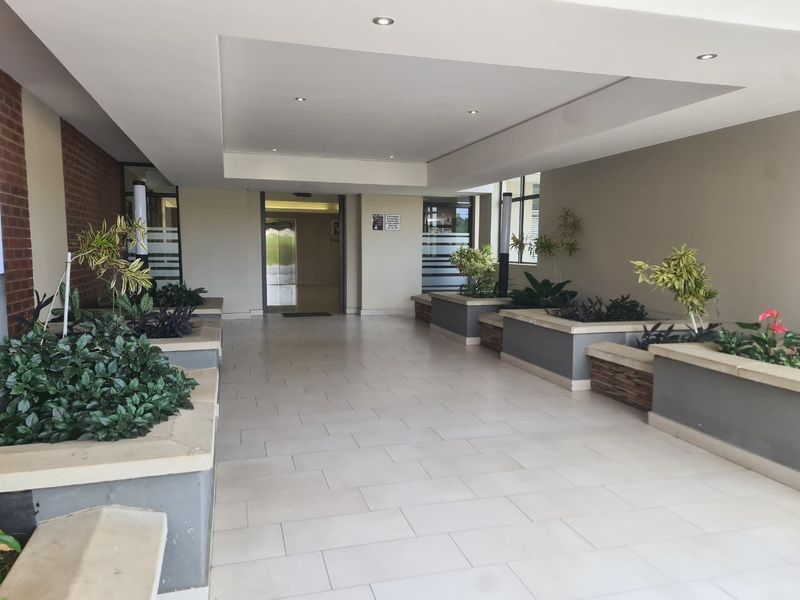 A spacious and well-maintained apartment building entrance lobby featuring tiled flooring, recessed ceiling lighting, and integrated built-in planters.