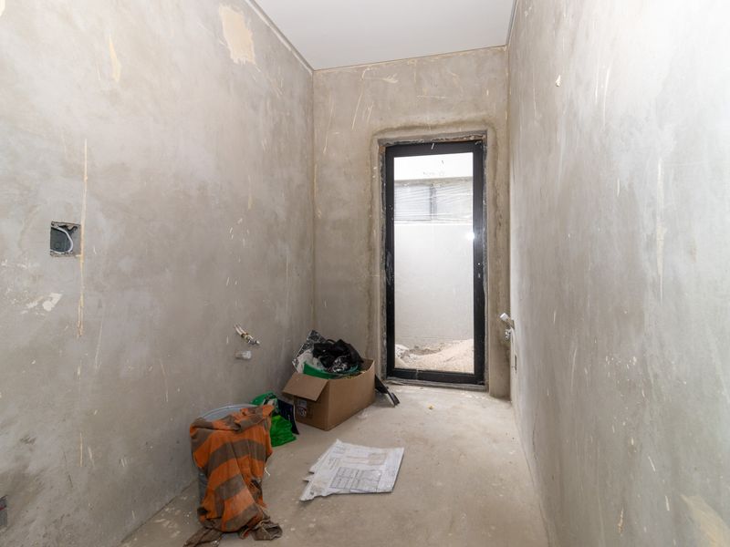 An unfinished utility or laundry space featuring exposed plaster walls, plumbing connections, and a modern aluminium framed door leading to the exterior.
