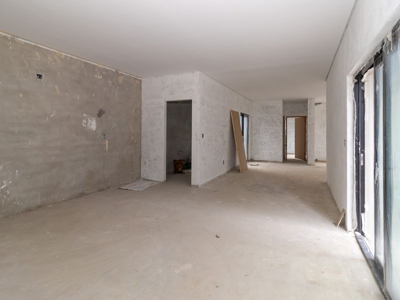 An unfinished open-plan living area featuring concrete floors, plastered walls, and large sliding glass doors, currently in a shell state awaiting final finishes.