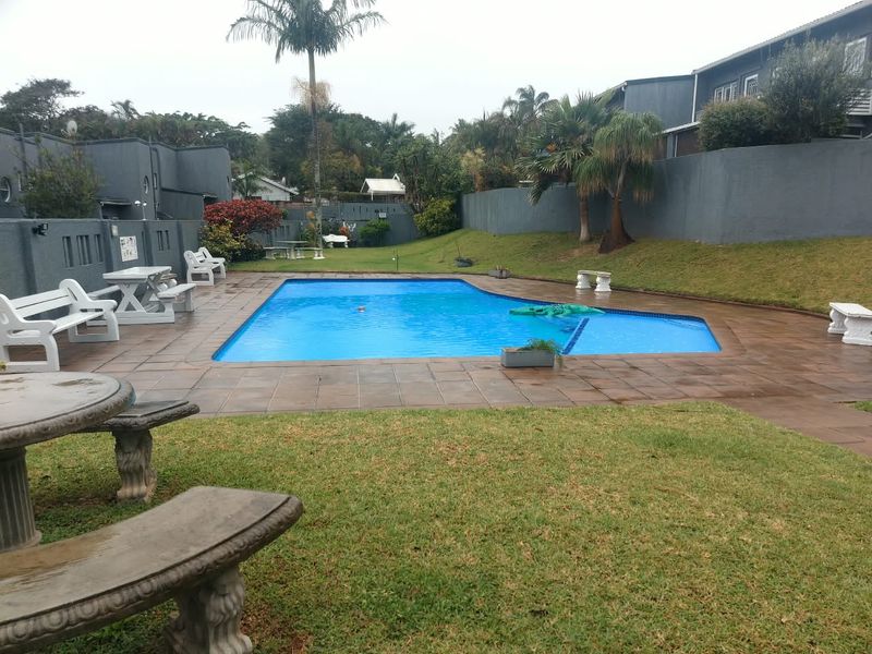 A communal swimming pool area featuring a paved deck, surrounding boundary walls, and landscaped garden space within the apartment complex.