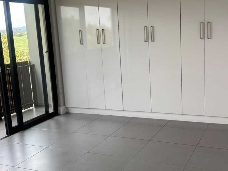A bright, modern bedroom featuring large built-in cupboards, sleek grey floor tiles, and a sliding glass door providing access to a private balcony.