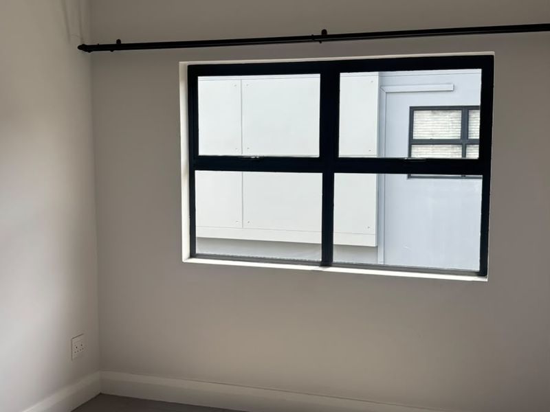 A bright, empty bedroom featuring neutral tiled flooring, modern black aluminium window frames, and recessed downlighting, offering a clean and well-maintained space.