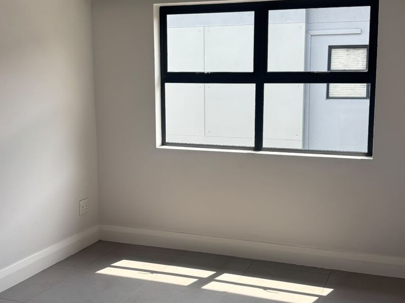 A bright, empty bedroom featuring modern grey tiled flooring, recessed downlights, and a black aluminium window frame.