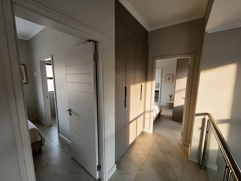 A bright, modern hallway featuring tiled flooring, built-in cupboards, and a stainless steel balustrade, providing access to multiple bedrooms.