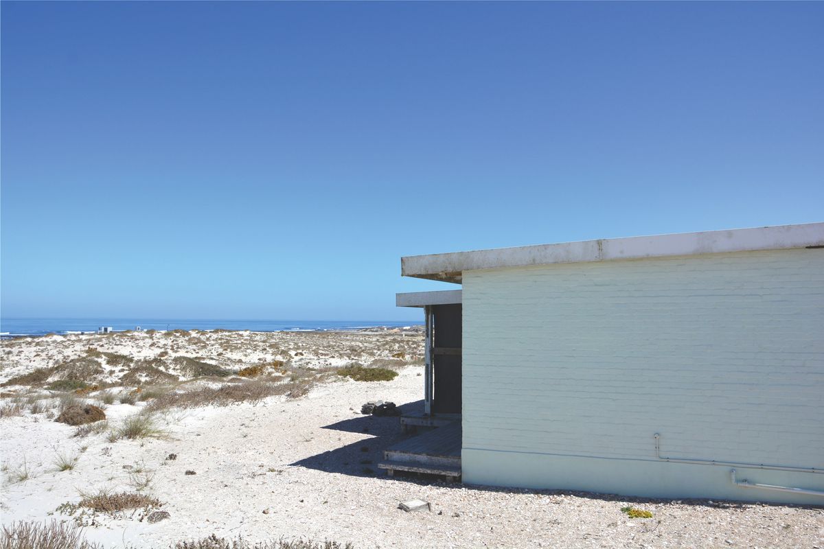 A view of the rear exterior of a coastal property, featuring painted brick walls and a flat roof, set against a natural sandy landscape.