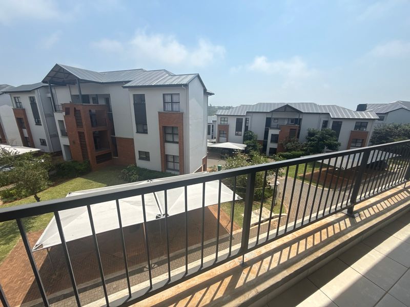 A spacious balcony with metal railings overlooking the apartment complex grounds, paved parking areas, and landscaped gardens.