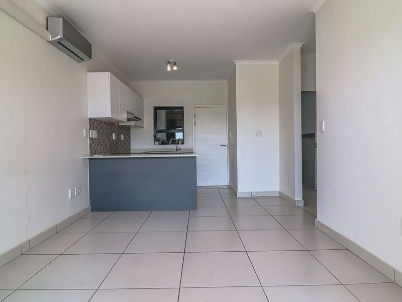 An open-plan living and kitchen area featuring neutral tiled flooring, modern cabinetry, a breakfast island, and a wall-mounted air conditioning unit.