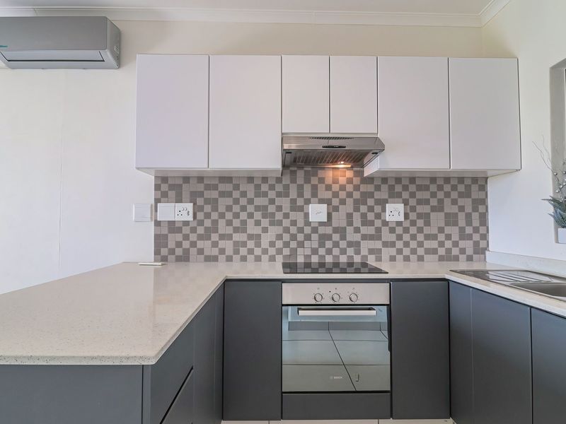 Modern kitchen featuring sleek grey and white cabinetry, stone countertops, mosaic backsplash, integrated appliances, and a wall-mounted air conditioning unit.
