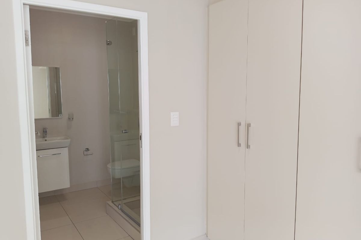 A clean, modern bedroom featuring built-in cupboards and an en-suite bathroom with a glass shower enclosure and tiled flooring.