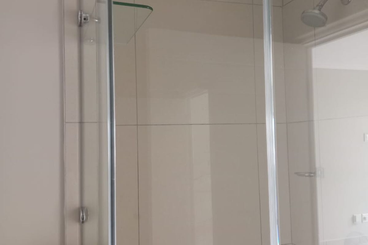 A clean, modern bathroom shower enclosure featuring neutral wall tiles, a fixed shower head, and a built-in glass shelf.
