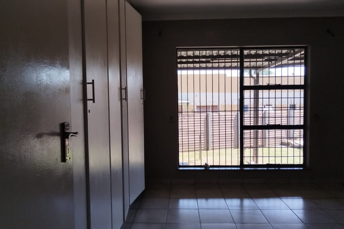 A spacious bedroom featuring tiled flooring, built-in cupboards, and a window fitted with security bars, offering a functional and secure living space.