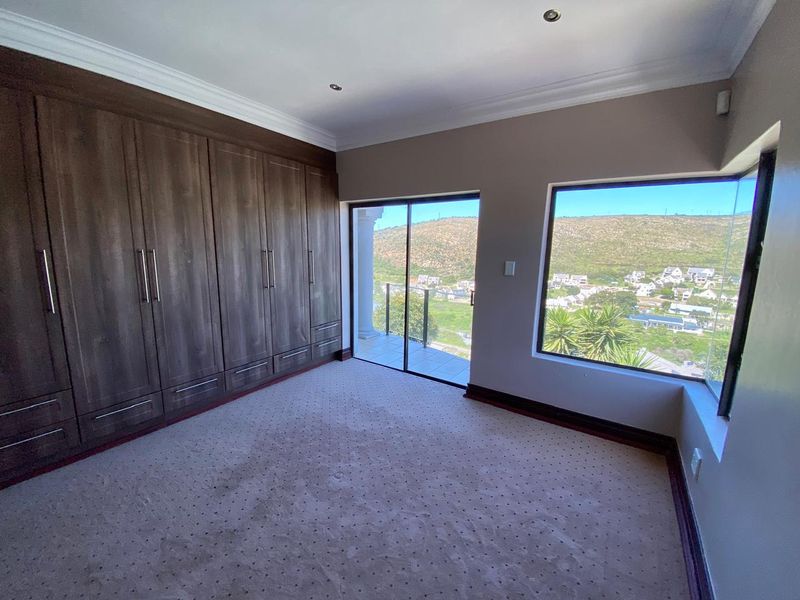 A spacious, empty bedroom featuring extensive built-in wooden cupboards, carpeted flooring, and a sliding door leading to a balcony with scenic hillside views.