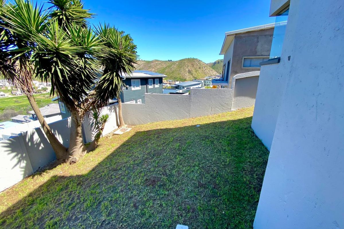 A rear garden area featuring a sloped lawn, boundary walls, and a glass balustrade, offering views of the surrounding neighborhood and hills.