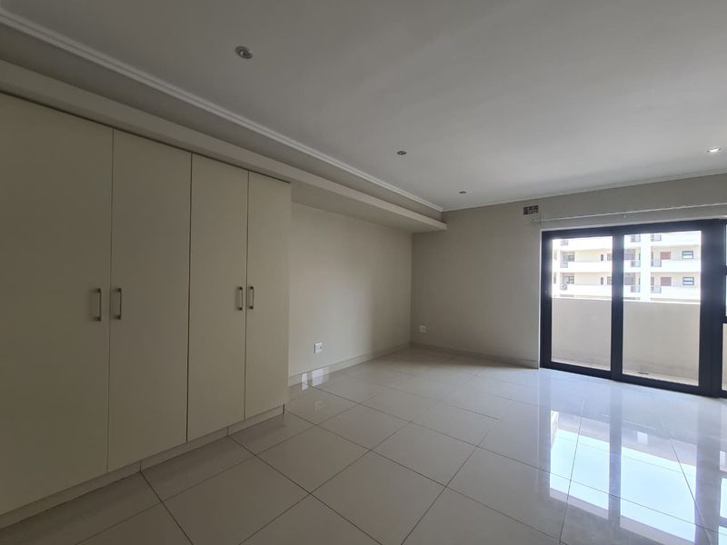 A bright, empty bedroom featuring neutral tiled flooring, built-in cupboards, and a sliding glass door leading to a balcony.
