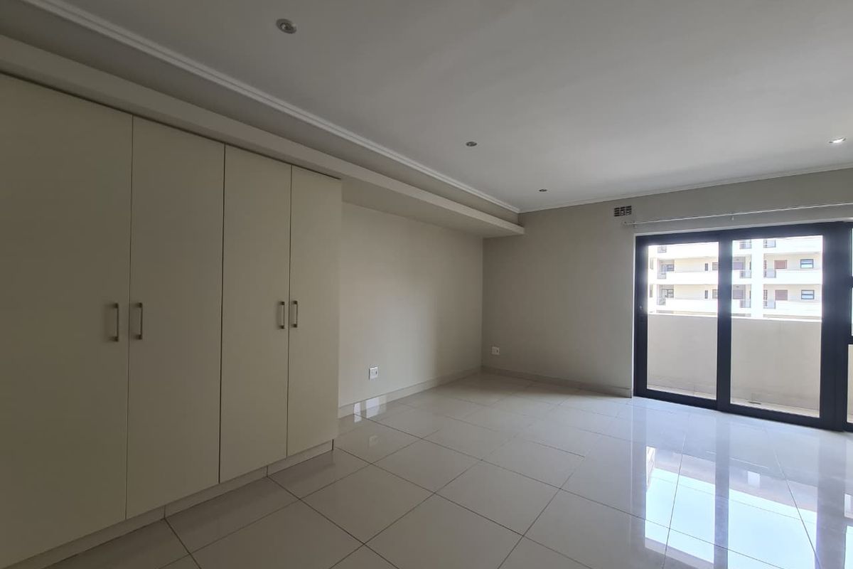A bright, empty bedroom featuring neutral tiled flooring, built-in cupboards, and a sliding glass door leading to a balcony.
