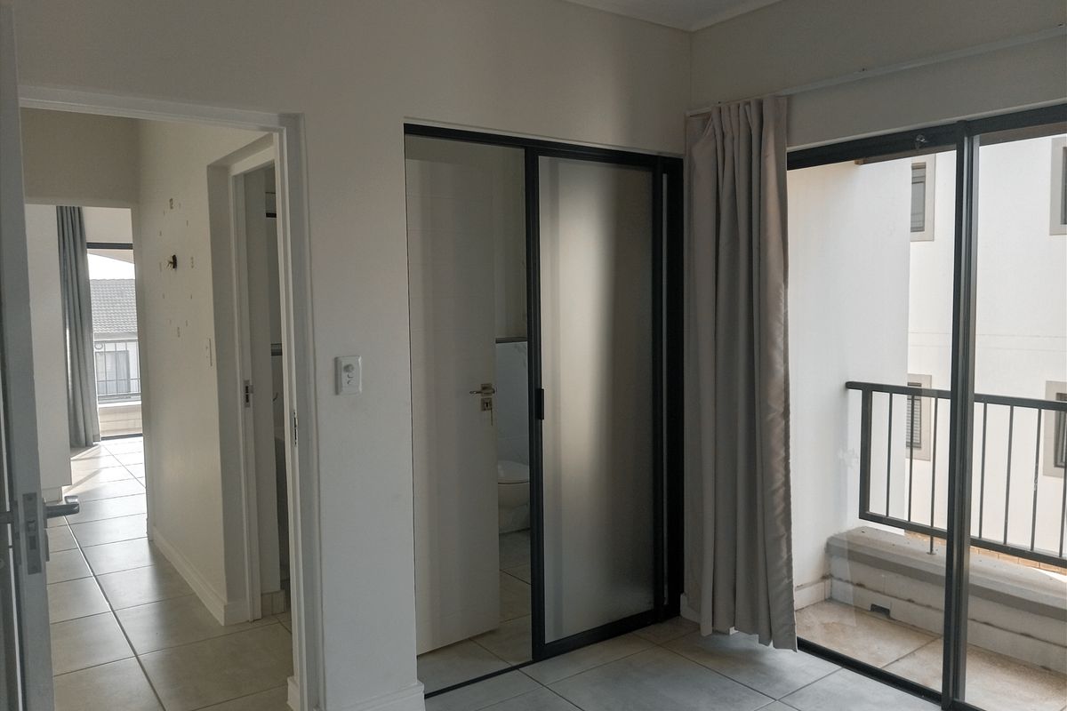 A bright, empty bedroom featuring tiled flooring, a sliding glass door leading to a private balcony, and an adjacent en-suite bathroom.