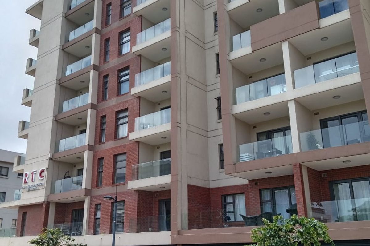 A modern multi-story apartment building featuring brick and concrete facade elements with glass-fronted balconies and paved street-level access.