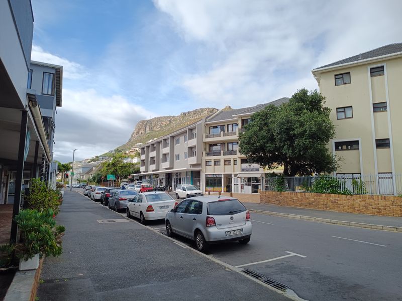 A street-level view of the apartment building exterior, showing the surrounding neighborhood, paved sidewalk, and road access.