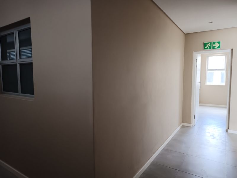 A clean, neutral-toned hallway featuring tiled flooring, aluminium window frames, and a clearly marked emergency exit route.