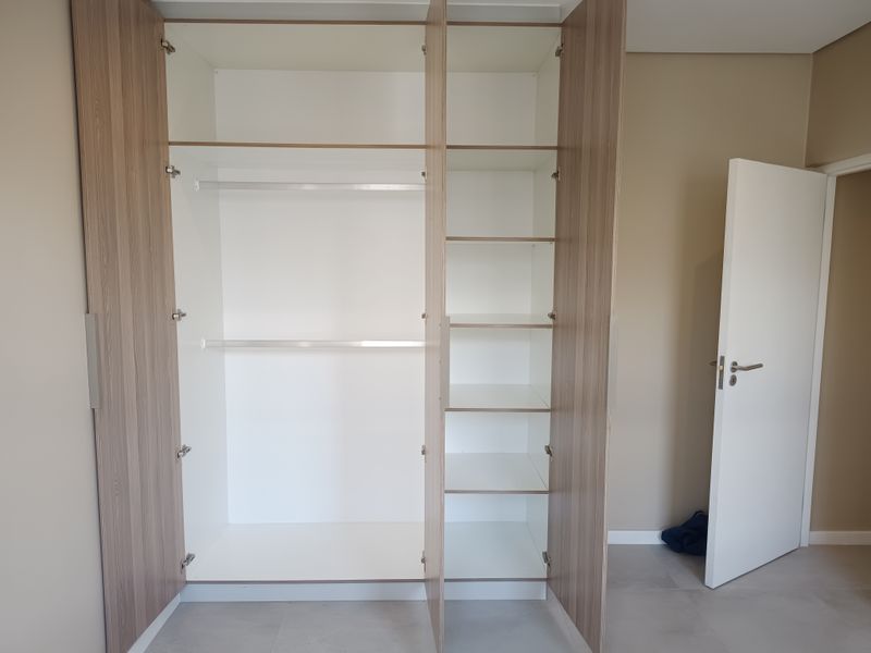 A clean, modern bedroom featuring a spacious built-in wardrobe with integrated shelving and hanging rails, finished in a light wood-grain laminate.