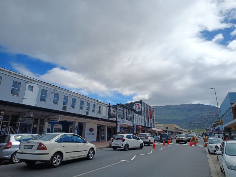 A street-level view of a commercial area featuring storefronts, paved roads, and mountain views in the background.