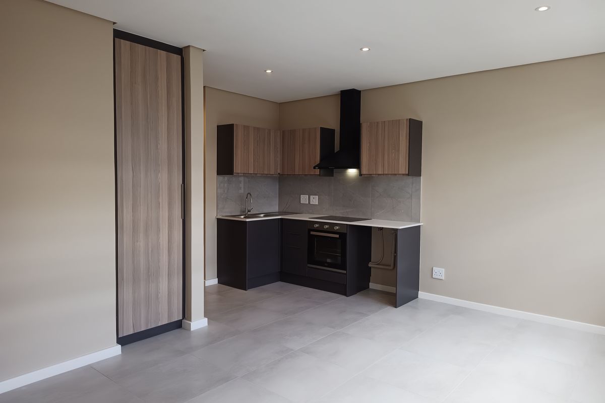 Modern open-plan kitchen featuring sleek dark cabinetry, stone countertops, integrated oven, extractor fan, and neutral tiled flooring in a well-lit, contemporary apartment space.