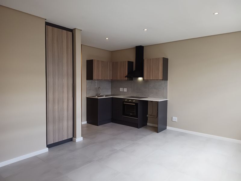 Modern open-plan kitchen featuring sleek dark cabinetry, stone countertops, integrated oven, extractor fan, and neutral tiled flooring in a well-lit, contemporary apartment space.