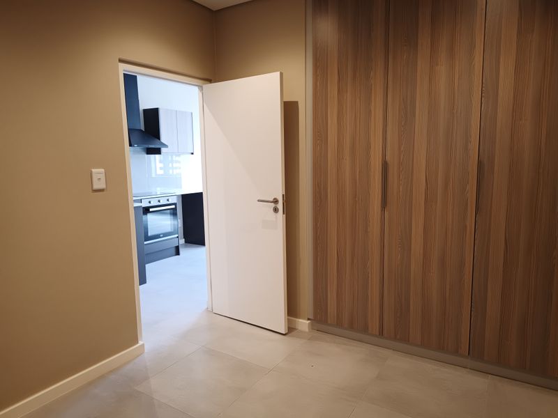 A clean, modern bedroom featuring built-in wood-grain cupboards, neutral wall tones, and tiled flooring, with a view into the adjacent kitchen area.