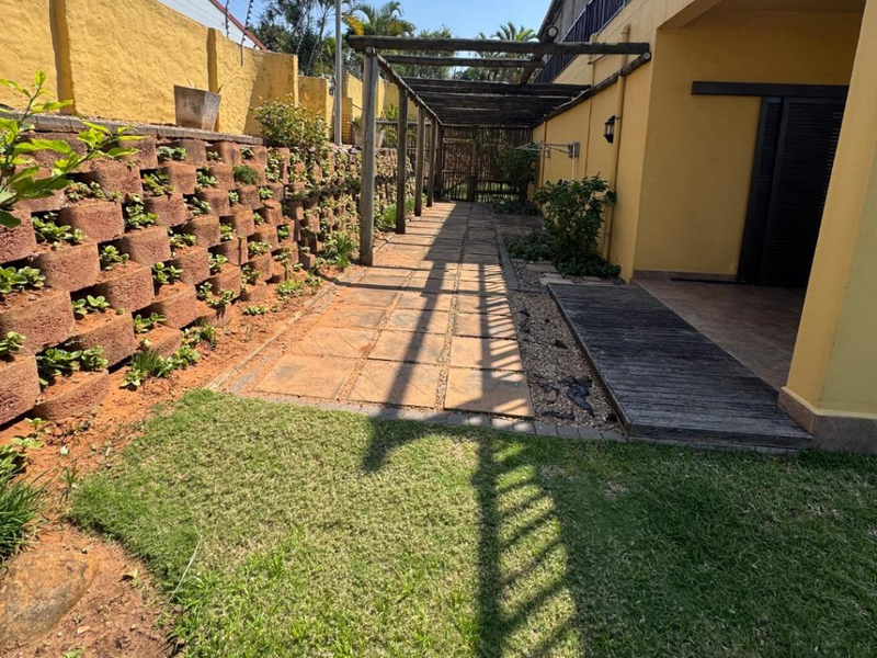 A rear exterior space featuring a paved walkway, timber pergola, and a tiered retaining wall with integrated garden beds.