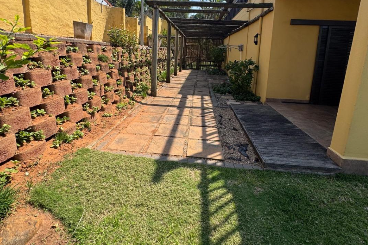 A rear exterior space featuring a paved walkway, timber pergola, and a tiered retaining wall with integrated garden beds.
