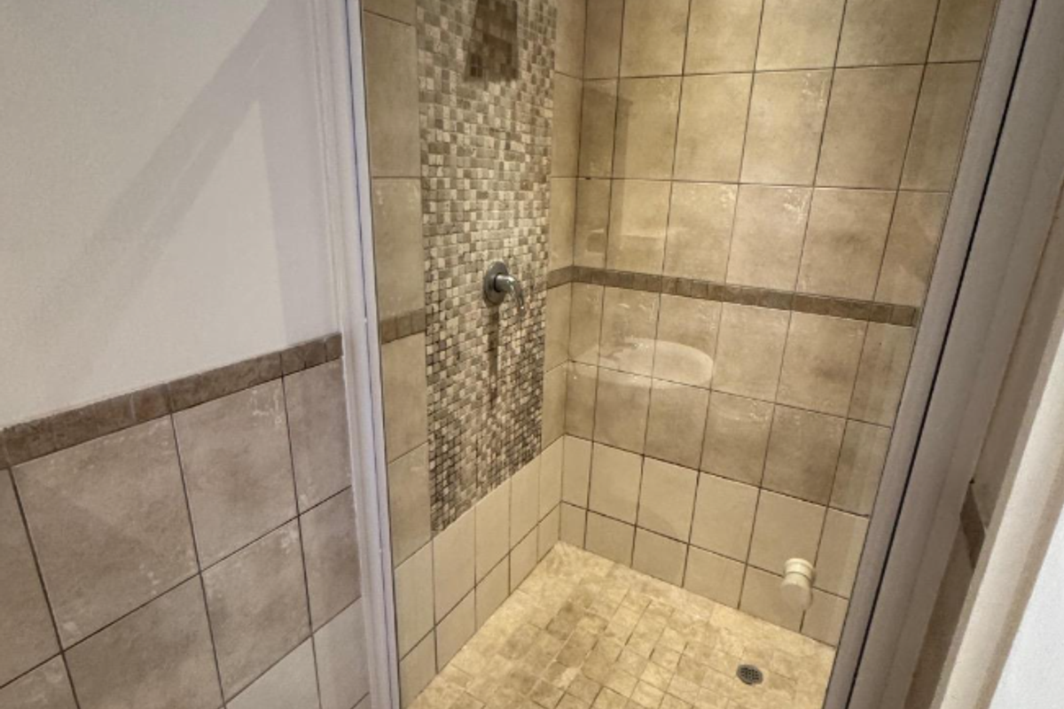 A functional shower stall featuring neutral-toned ceramic wall tiles with a decorative mosaic accent strip and a white-framed glass enclosure.
