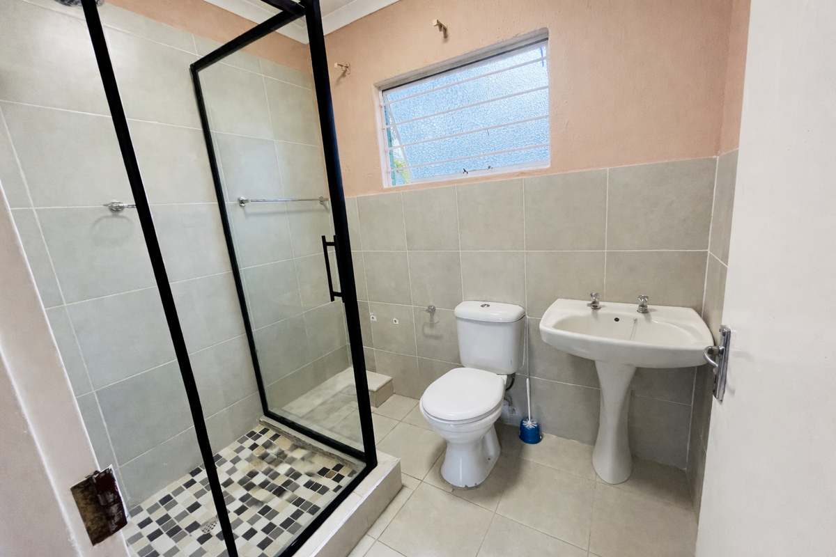 A functional bathroom featuring a glass-enclosed shower, pedestal basin, and toilet, with neutral wall and floor tiling throughout.