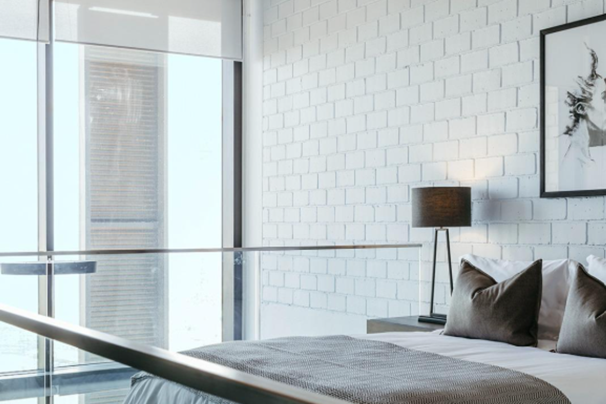 Modern bedroom featuring an exposed concrete ceiling, painted brick feature wall, and floor-to-ceiling windows with a glass balustrade overlooking the space.