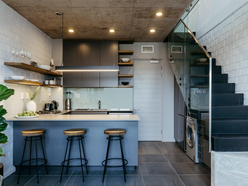 Modern open-plan kitchen featuring sleek cabinetry, stone countertops, exposed concrete ceiling, and a striking glass-railed staircase in a contemporary industrial-style apartment.