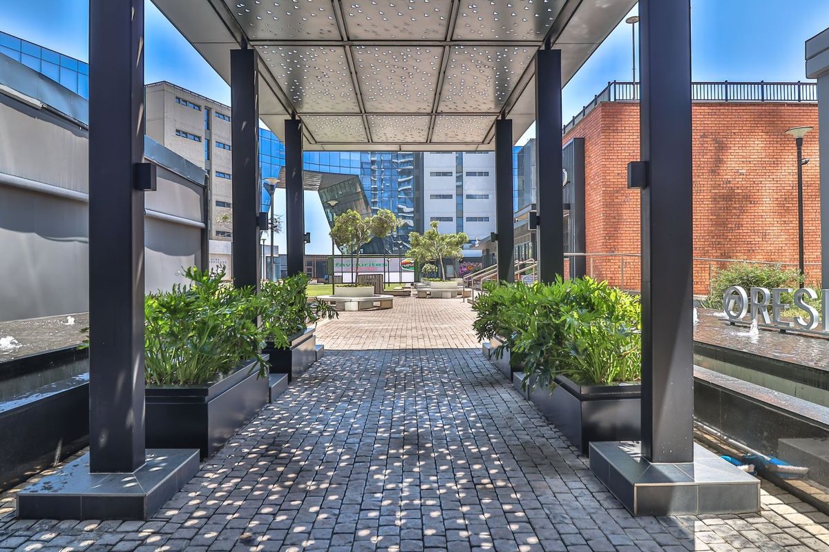 A modern outdoor courtyard featuring a perforated metal canopy, cobblestone paving, built-in planters, and a decorative water feature.
