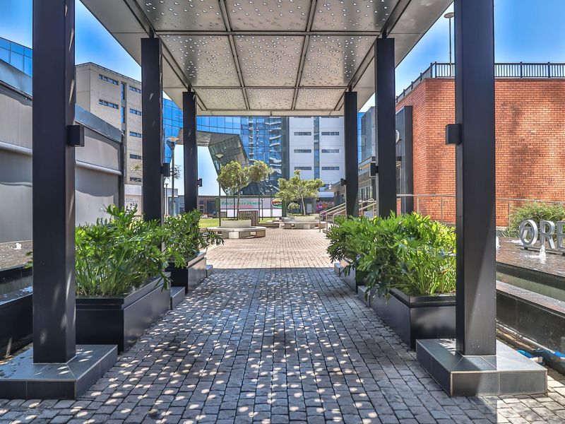 A modern outdoor courtyard featuring a perforated metal canopy, cobblestone paving, built-in planters, and a decorative water feature.
