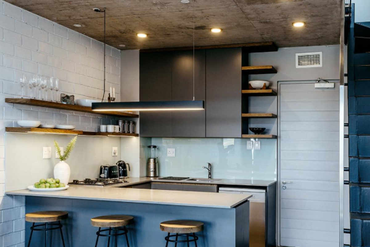 Modern kitchen featuring an exposed concrete ceiling, painted brick walls, sleek cabinetry, stone countertops, and integrated appliances with a breakfast bar.