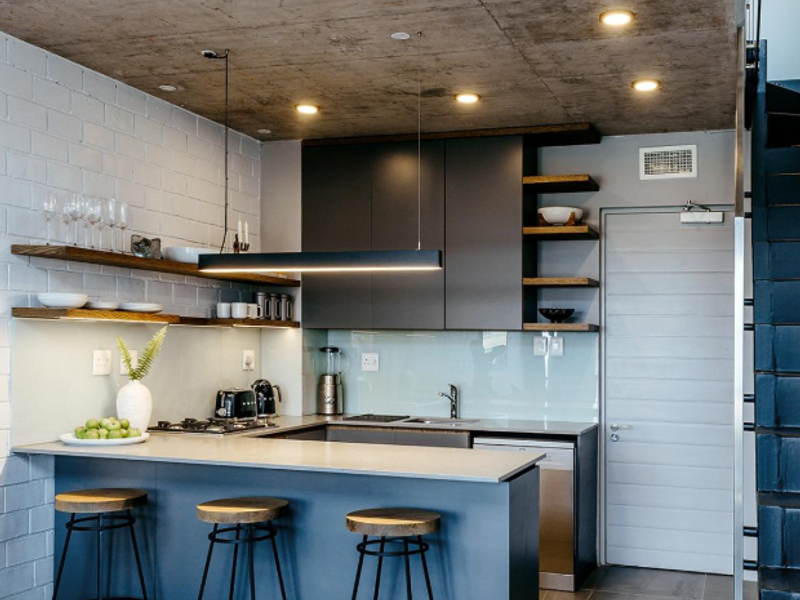 Modern kitchen featuring an exposed concrete ceiling, painted brick walls, sleek cabinetry, stone countertops, and integrated appliances with a breakfast bar.